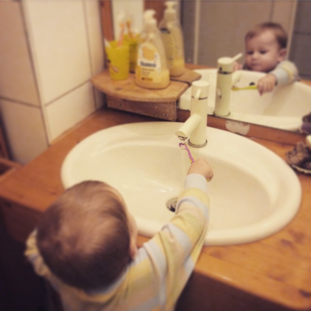 Das Kind steht auf einem Hocker  und hält seine Zahnbürste unter das fließende Wasser am Waschbecken.